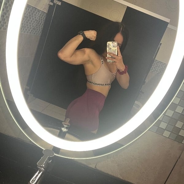 Young woman flexing her toned arms in a gym mirror selfie, wearing a sports bra and leggings.
