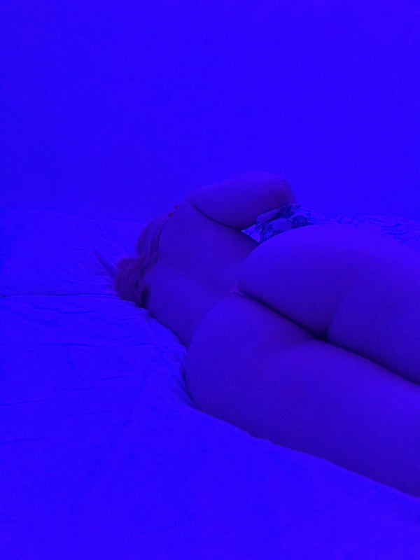 A person lying on a bed in a dimly lit room with blue lighting, viewed from behind.