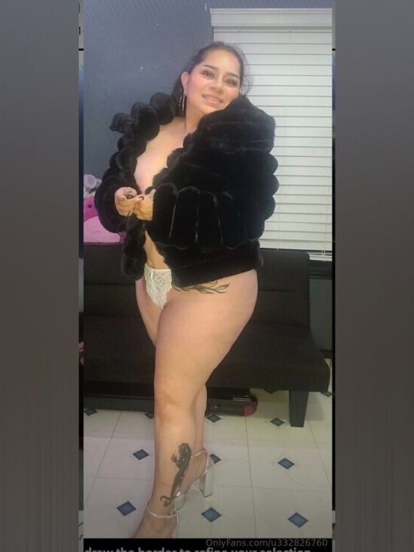 Woman standing in a fluffy black jacket and light bottoms, posing confidently indoors with tiled floor.