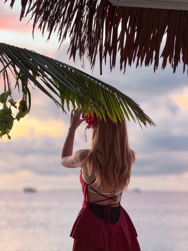 A woman standing on a beach at sunset, wearing a red bikini and holding a hibiscus flower under a thatched roof with palm leaves.