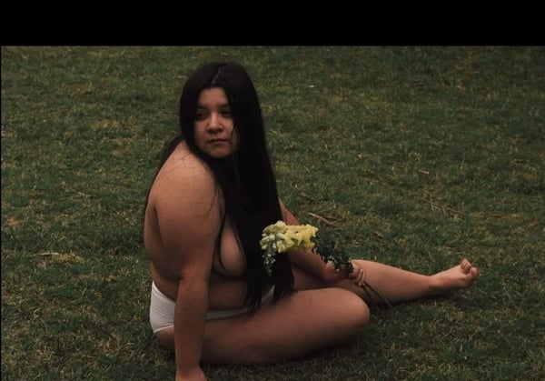 A woman sitting on grass holding yellow flowers, dressed in underwear with a scenic outdoor background.