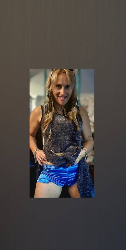 A woman with long wavy blonde hair posing confidently indoors, wearing a dark tank top and blue shorts.