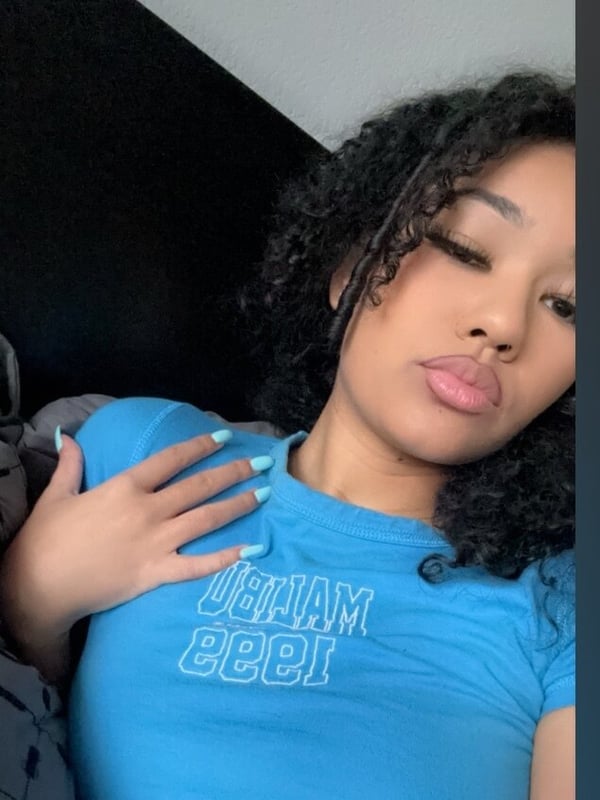 Close-up of a young woman with curly black hair, wearing a blue graphic t-shirt, posing with her hand on her shoulder.