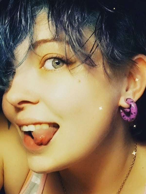 A woman with dyed blue hair and large purple hoop earrings, smiling playfully with her tongue out, wearing a pink necklace against a dark starry background.
