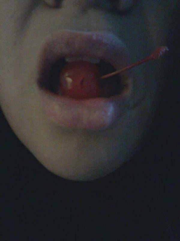 Close-up portrait of a woman with glossy lips holding a red cherry in her mouth against a dark background.