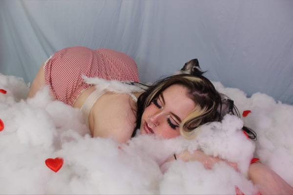 Young woman with cat ears and colorful hair lounging on a bed of white clouds surrounded by red hearts, wearing a checkered outfit.