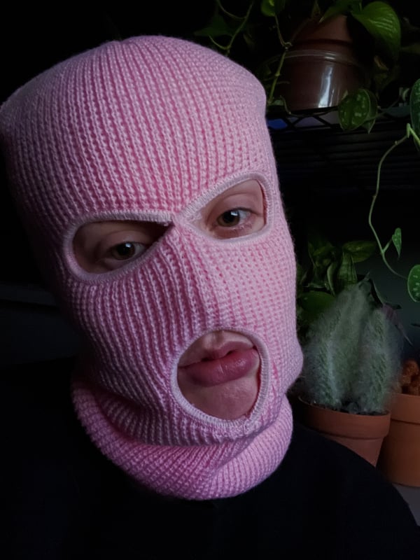 Close-up of a person wearing a pink balaclava mask with eye and mouth openings, set against a dark background with plants.