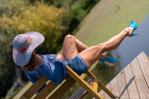 Woman lounging on a wooden dock by the water, wearing a colorful hat, blue top, denim shorts, and high heels, with a scenic outdoor background.