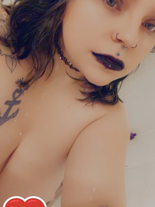 Close-up selfie of a woman with dark wavy hair, heavy gothic makeup, choker necklace, and visible tattoos and piercings, smiling playfully.