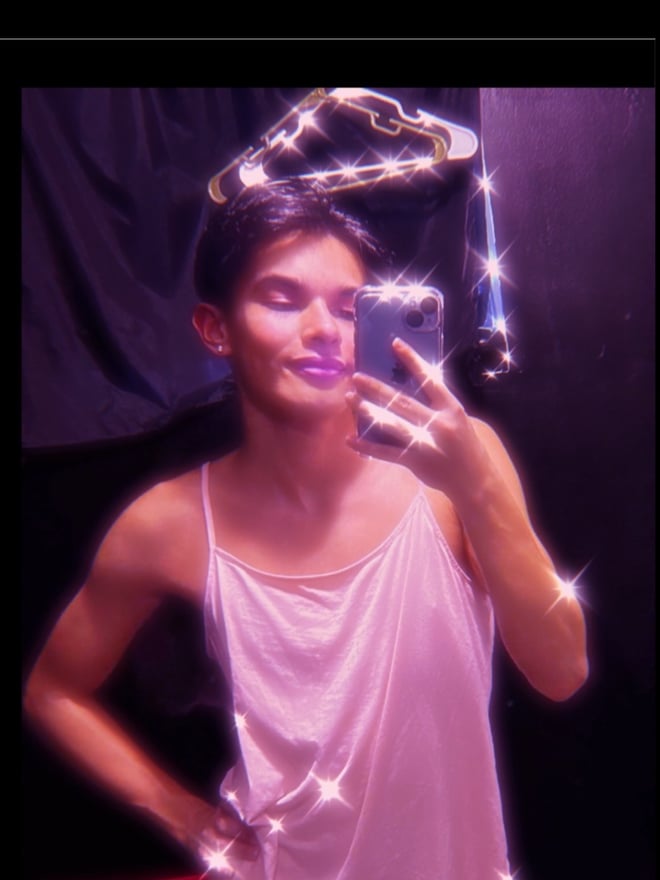 Person taking a selfie in a pink tank top with glitter effects against a dark background.