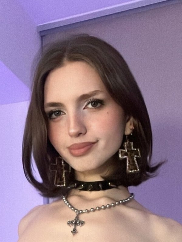 A close-up portrait of a person with shoulder-length brown hair, wearing cross-shaped earrings and a choker necklace.