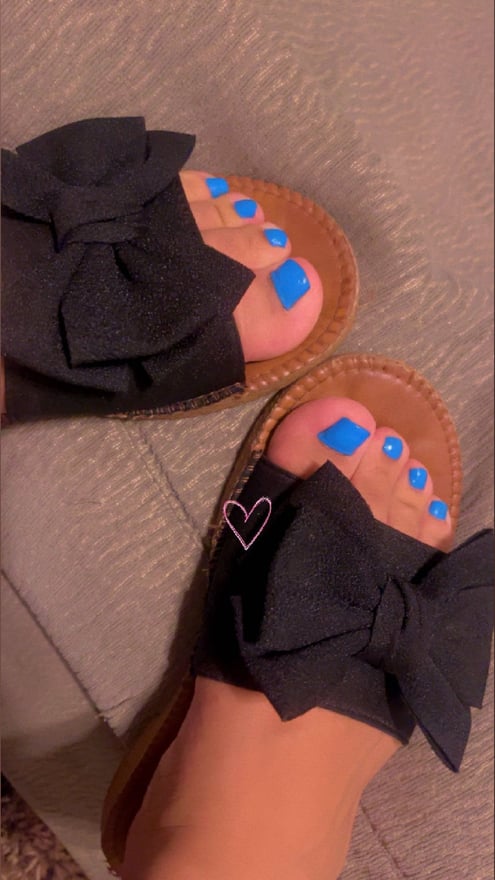 Close-up view of a person's feet wearing black bow sandals with blue painted toenails on a neutral background.
