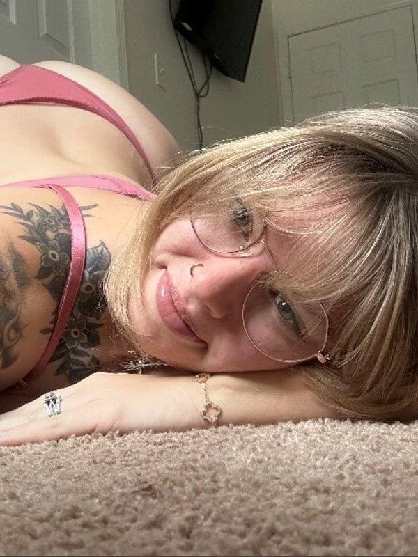 Woman with blonde hair and glasses lying on a bed in pink lingerie, smiling at the camera with tattoos visible on her arm.