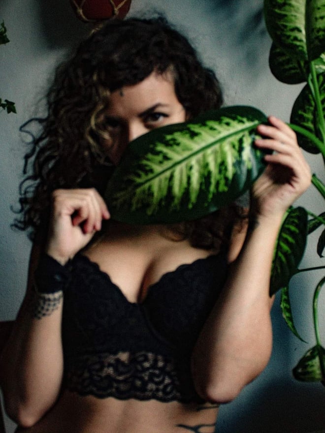 A woman with curly hair holds a large green leaf in front of her face, wearing black lace top, surrounded by houseplants.