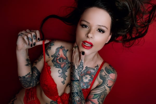 A woman with long dark hair and extensive tattoos poses playfully on a red background, wearing red lingerie and dark nail polish.