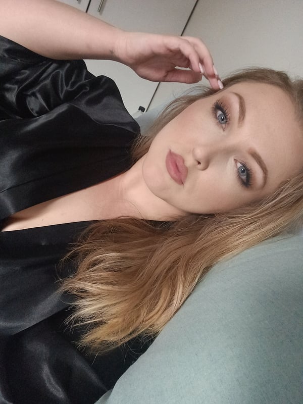 A woman with long blonde hair wearing a black satin top, posing with hand on face in a casual indoor setting.