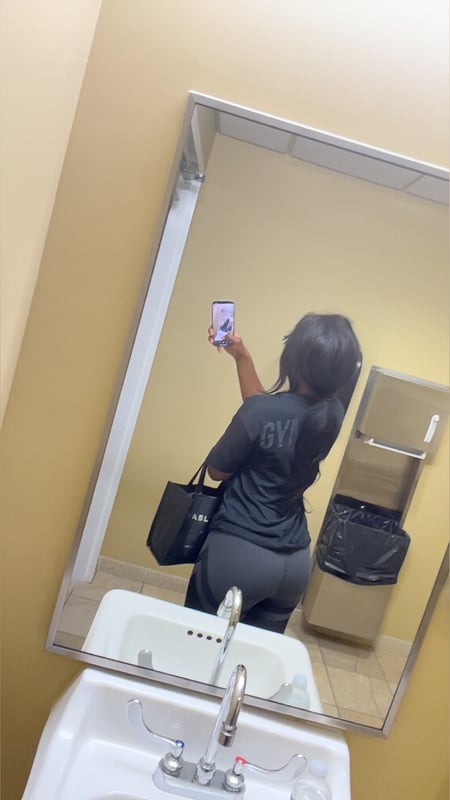A young woman with long dark hair stands in front of a mirror in a bathroom, wearing a gray shirt and pants while holding her phone.