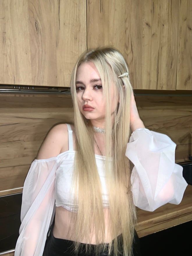 A young woman with long blonde hair stands in front of wooden cabinets, wearing a white off-shoulder top with sheer sleeves and black pants, gazing at the camera.