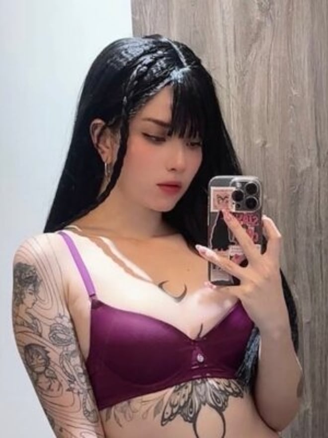 A woman with long black hair takes a mirror selfie while wearing dark lingerie, with visible tattoos on her arm and torso.