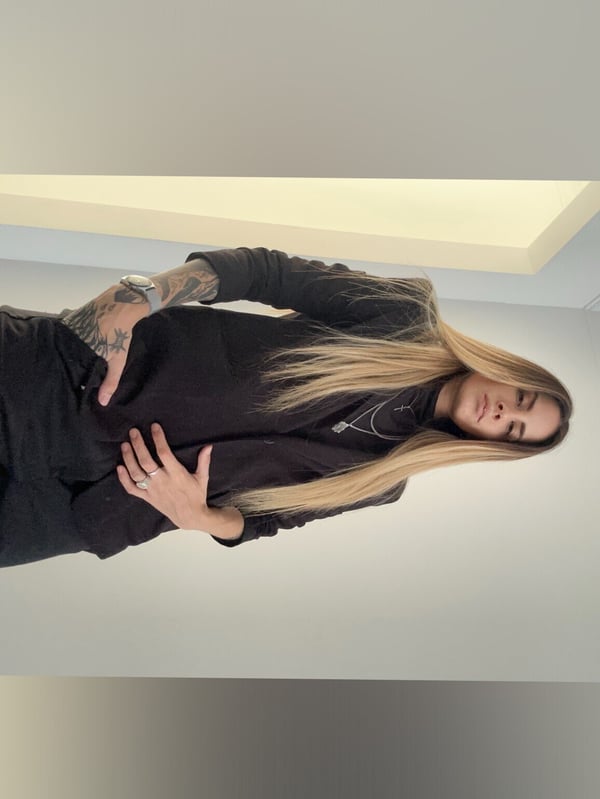 A woman with long blonde hair posing confidently in a black long-sleeve top and pants, hands on hips, against a plain background with soft lighting.