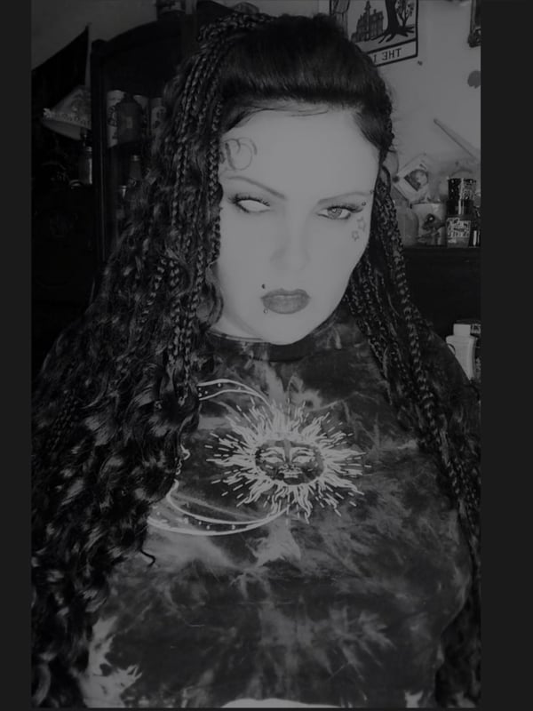 Woman with long braided black hair wearing a dark tie-dye shirt with a sun graphic, posing in a dimly lit room with gothic decor.