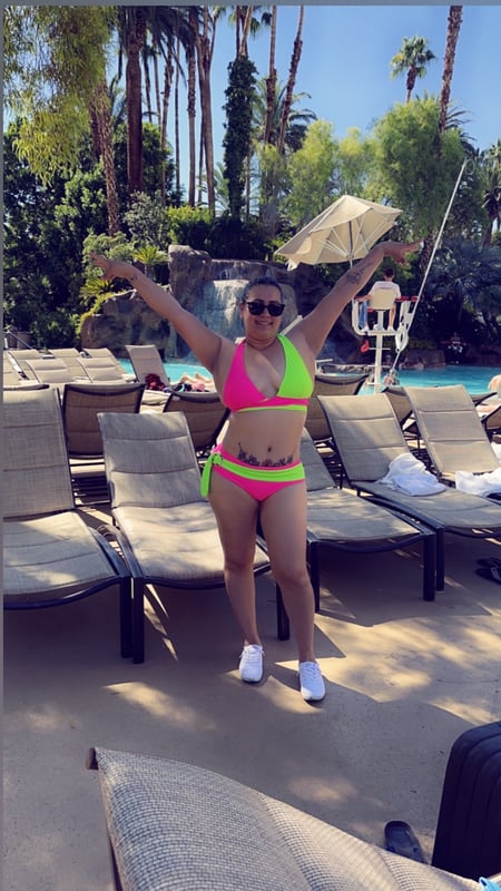 A woman in a bright pink bikini poses with arms raised at a resort pool area, holding a parasol, surrounded by palm trees and lounge chairs.