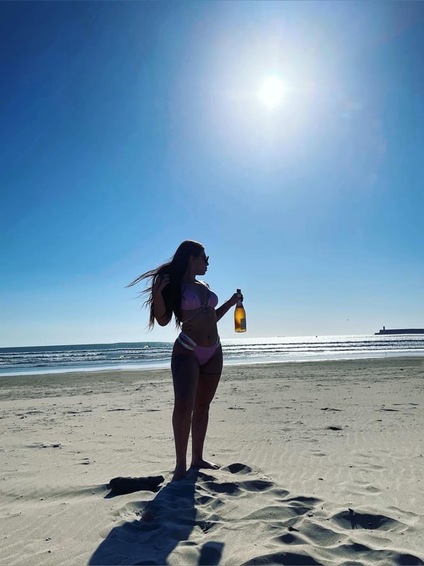 Young woman in a pink bikini standing on a sunny beach with ocean waves in the background, holding a bottle and gazing to the side.