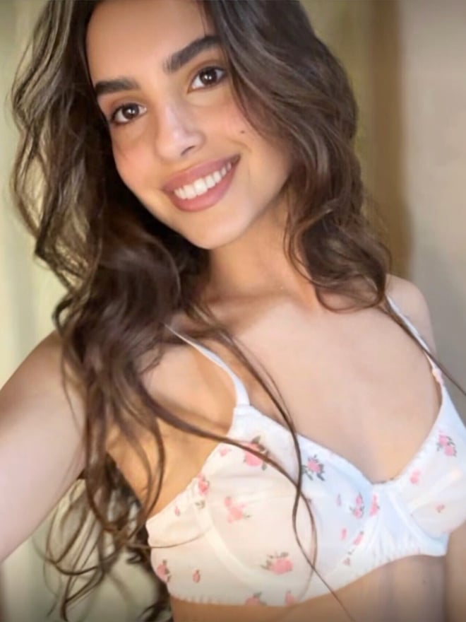 Young woman with long wavy hair smiling in a close-up selfie, wearing a light-colored top against an indoor background.