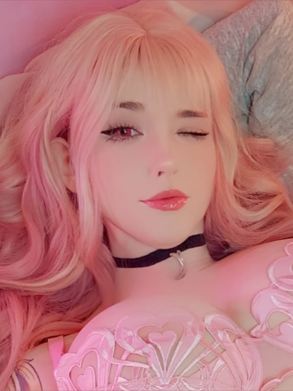 A person with long pink hair lying on a bed surrounded by pink pillows and plush toys, wearing a choker and lace outfit.