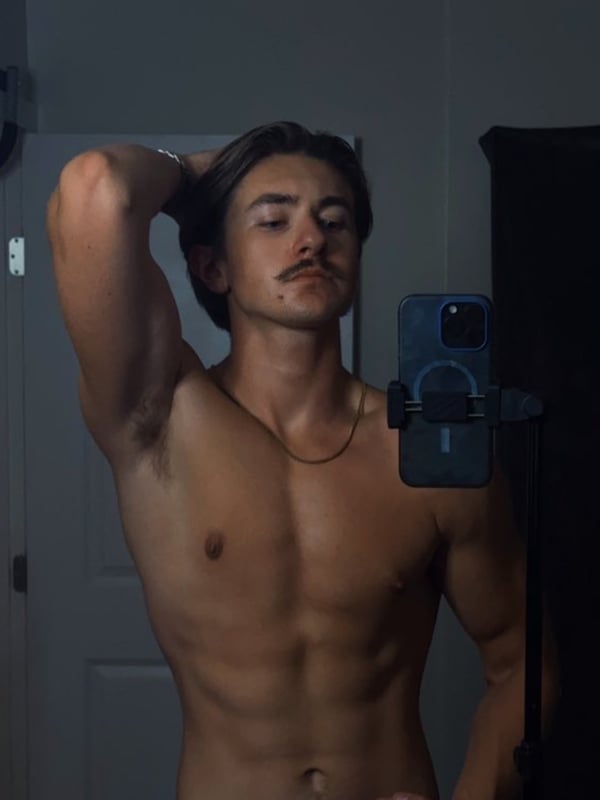 Young man with long hair posing shirtless in front of a mirror, flexing his arm while holding a phone.