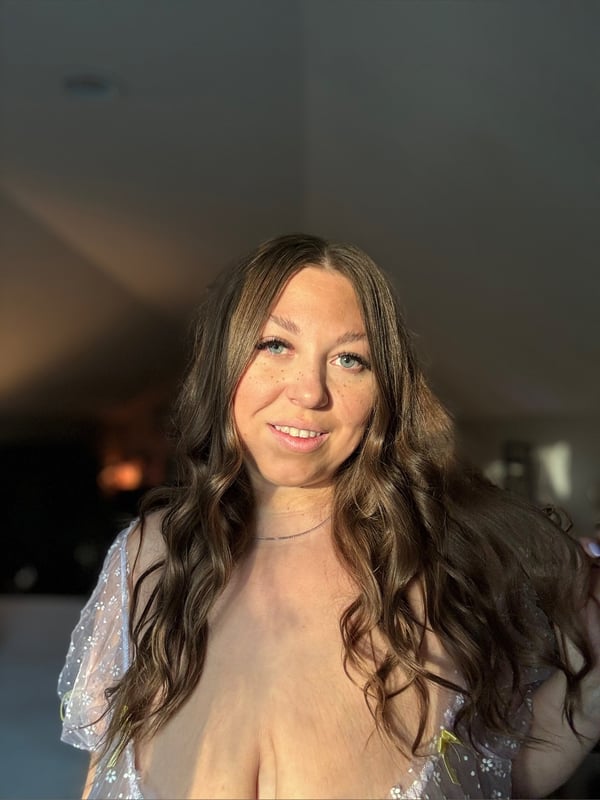 Woman with long wavy brown hair smiling warmly in a sheer white top, standing in a softly lit indoor setting.