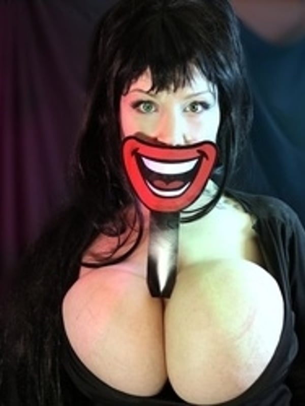 A woman with long dark hair holding a large red smiling mask in front of her face, dressed in black attire against a dark draped background.
