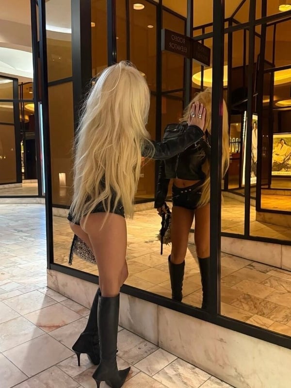A blonde woman in a black leather jacket, shorts, and high boots posing confidently in front of mirrored doors in a luxurious interior.