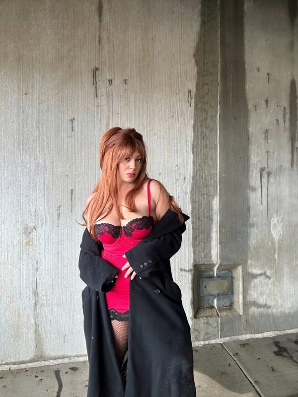Woman with red hair posing in red lingerie and a black coat against a concrete wall background.