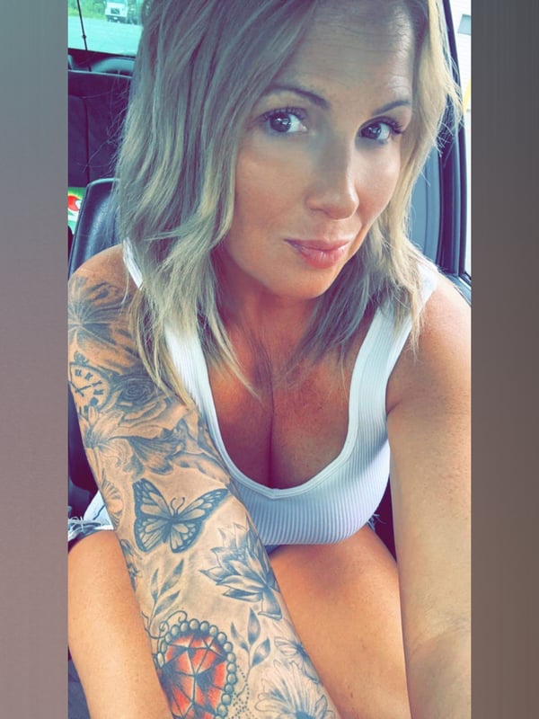 A woman smiling in a car, wearing a white tank top, with detailed arm tattoos visible.