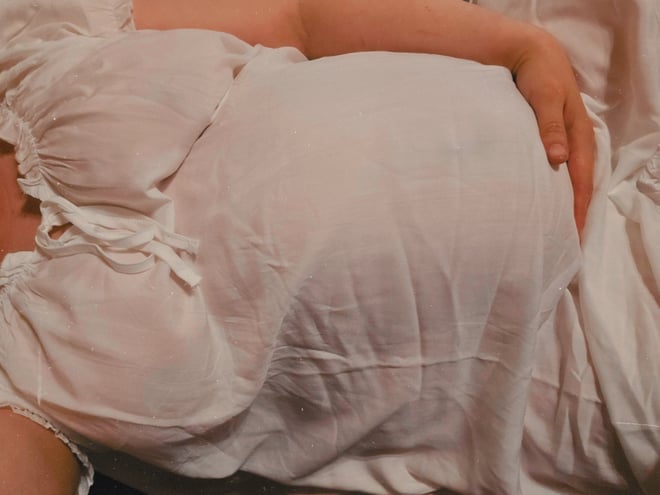 A woman in a sheer white lace top posing on a bed with pillows in a softly lit room.