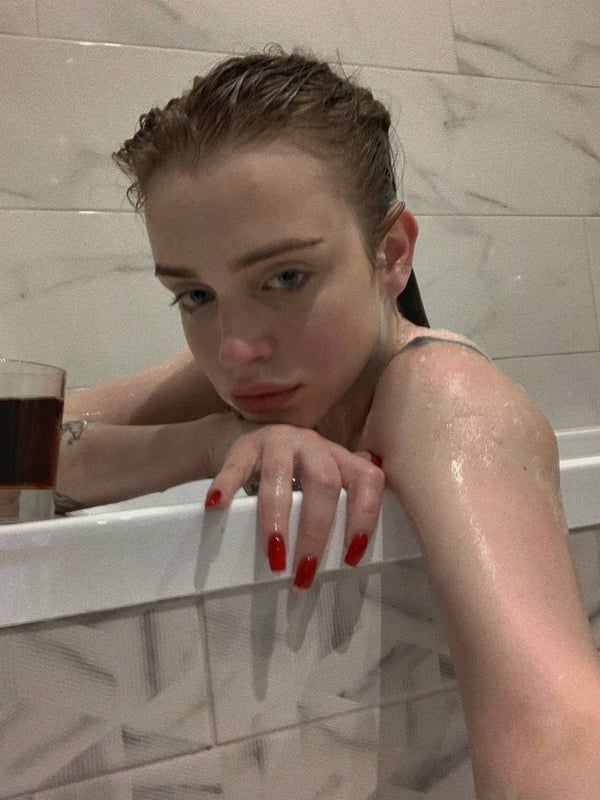 Young woman with wet hair leaning over the edge of a bathtub, holding a glass, with red nail polish.