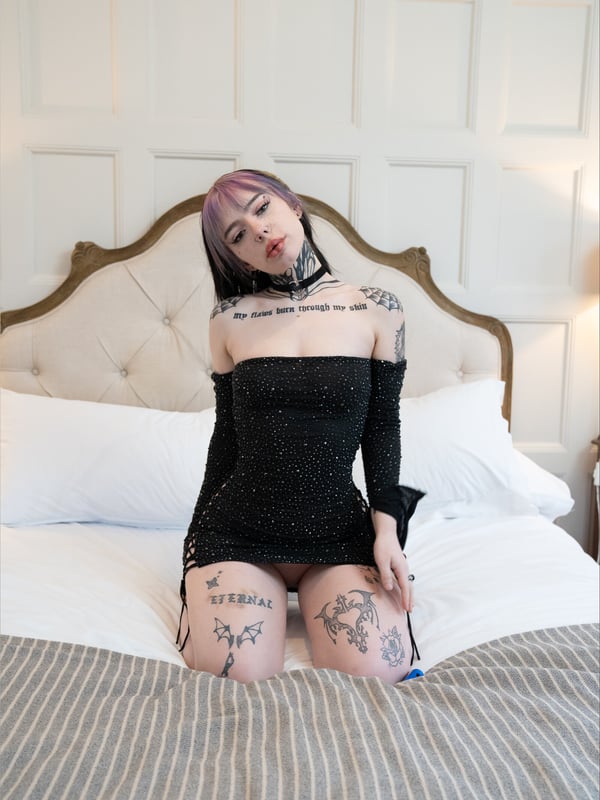 A young woman with dyed purple hair and visible tattoos sitting on a bed in a black sparkly dress, wearing a choker necklace.