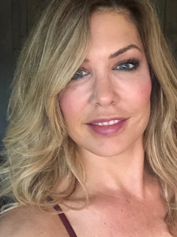Close-up portrait of a smiling woman with blonde wavy hair, wearing a lace camisole top, posed indoors against a rustic wooden background.