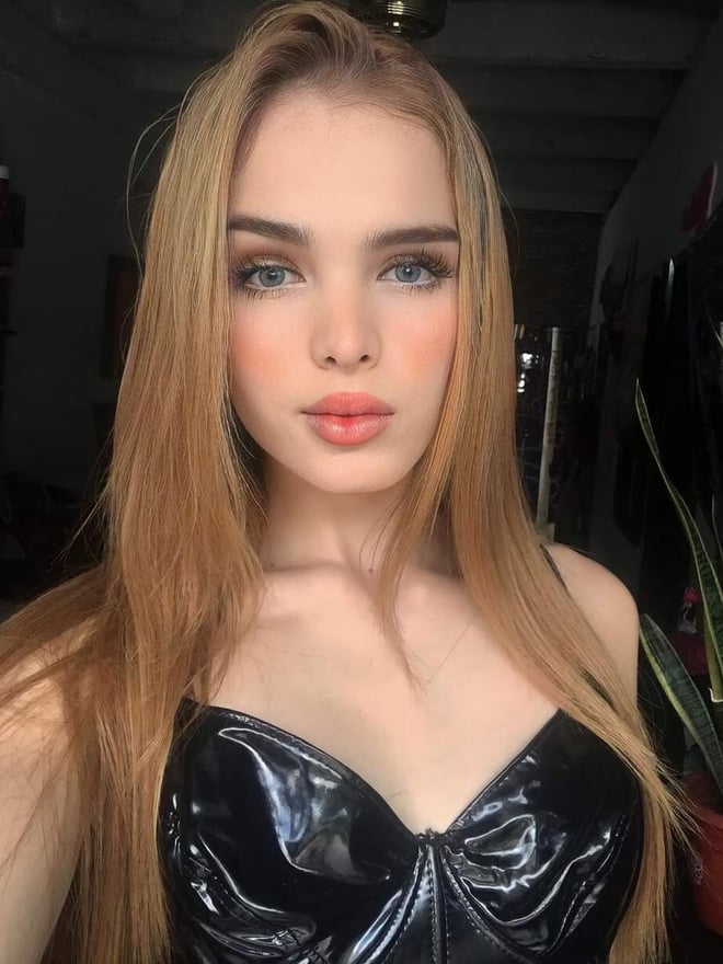 A young woman with long light-brown hair posing for a close-up portrait indoors, wearing a glossy black top.