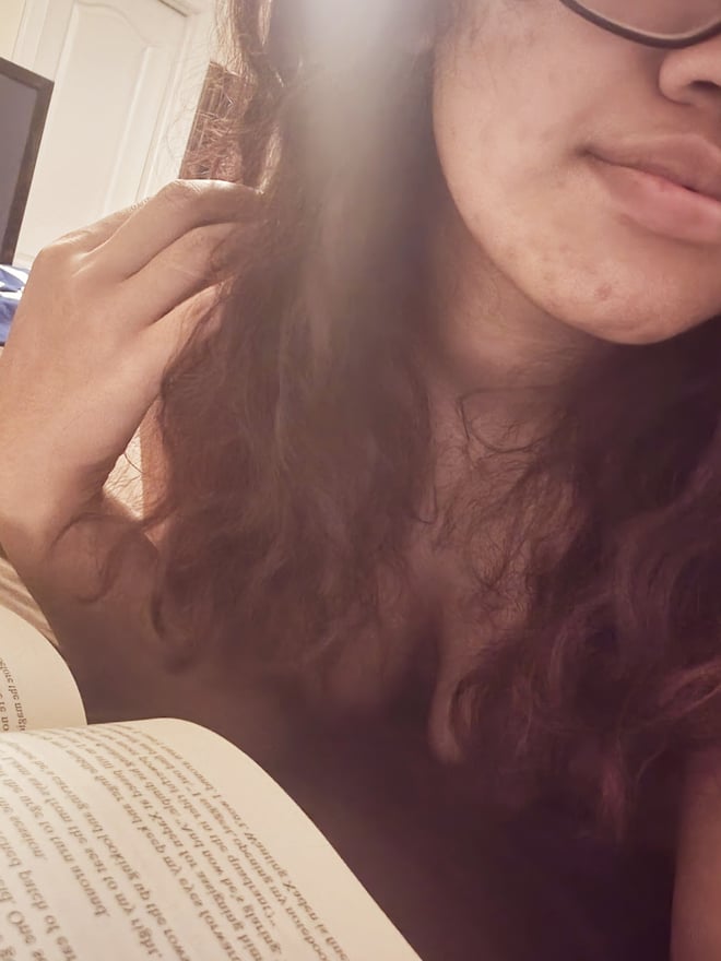 Close-up of a woman with curly hair and glasses, smiling softly while holding an open book indoors.