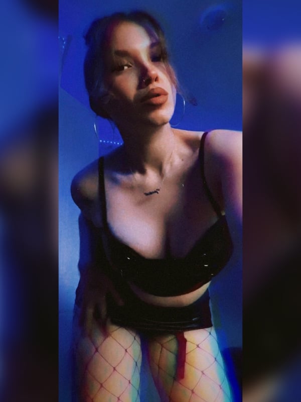 A woman with long hair posing confidently in a black top and fishnet stockings under blue lighting with bubbles in the background.