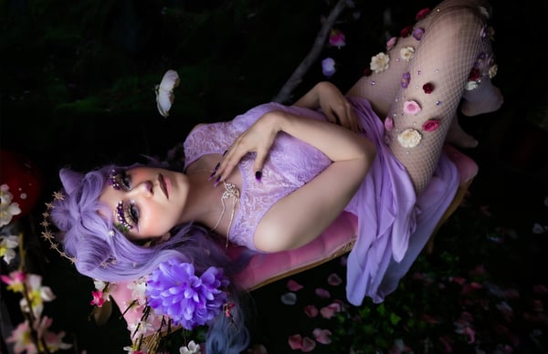 A person in an elaborate purple fantasy costume with floral elements and sheer stockings, reclining on a pink surface amid a dark, flower-strewn background.