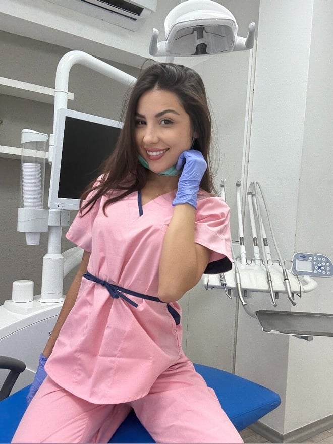 A smiling woman wearing pink scrubs and blue gloves, sitting in a dental office chair surrounded by dental tools.