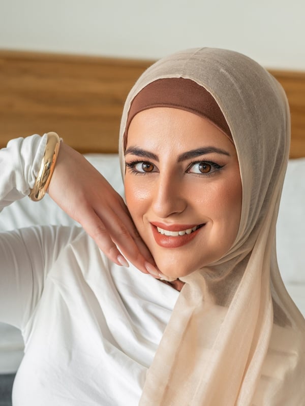 Close-up portrait of a smiling woman wearing a beige hijab and white top, posing with her hand on her cheek against a bedroom background.