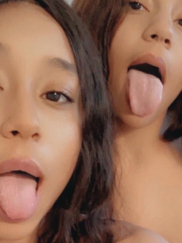 Two young women with long dark hair posing closely together, playfully sticking out their tongues.
