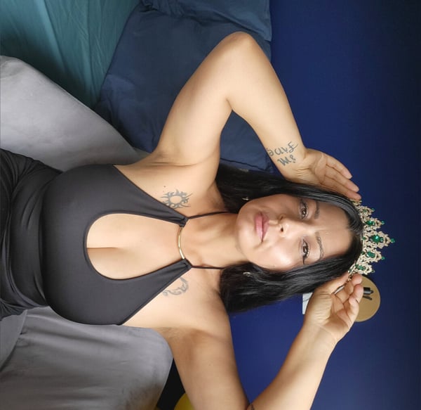 A woman with long dark hair posing on a bed while wearing a crown and a black top.