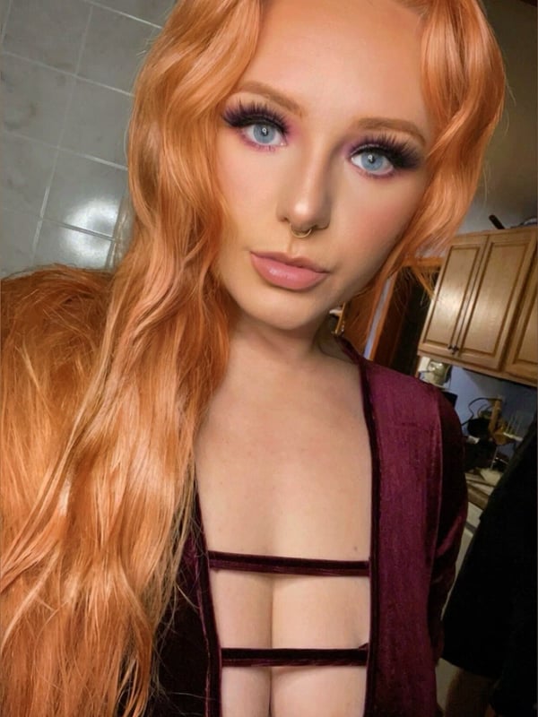 Young woman with long wavy orange hair posing confidently in a maroon top with cutouts, standing in a kitchen area with tiled walls.