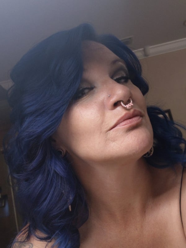 Close-up portrait of a woman with vibrant blue wavy hair, facial piercings, and tattoos, smiling confidently indoors.