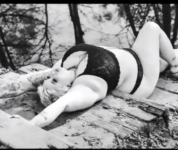 A woman with tattoos poses relaxed on a wooden surface in a forested area, wearing black lace lingerie.